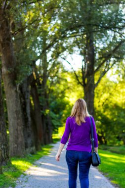 Yazın yaşam tarzının özünü yansıtan keyifli bir dikey görüntü. Sarışın bir kadın, arka plandan bakıldığında parkta rahatça yürürken mutluluk ve neşeyi somutlaştırır..