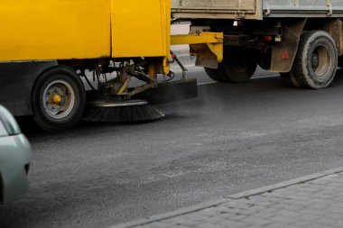Sarı bir yol temizleme makinesi bir şehir caddesindeki asfaltı temizliyor. Toz ve enkazı süpürürken dairesel fırça görünür..