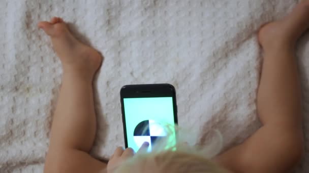 Ett porträtt av en söt baby kontrollera smartphone ligger i en säng — Stockvideo