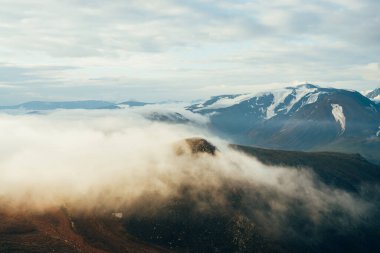 Atmosferik alp manzarası. Kayaların üzerinde parlak, altın gibi alçak bulutlar ve gün doğumunda buzulları olan dev karlı dağlar. Bulutların üzerinde altın saatte uçuyor. Muhteşem dağlık manzara.