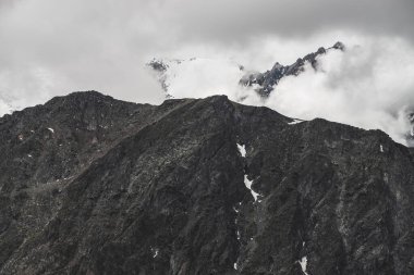 Atmosferik minimalist dağ manzarası. Büyük kayalık bir dağ duvarı ve alçak bulutlarda karla kaplı bir tepe. Devasa buzullu büyük dağlar. Karlı, kayalık bir duvar. Yüksek irtifada görkemli manzara.