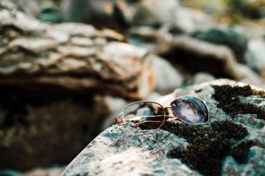 Broken sunglasses on stone with moss in sunlight. Lost thing on mossy boulder in sunny day. Glare on shiny thing. Nonsense happened. Lost sunglasses on rock close-up. Things break, life goes on. clipart