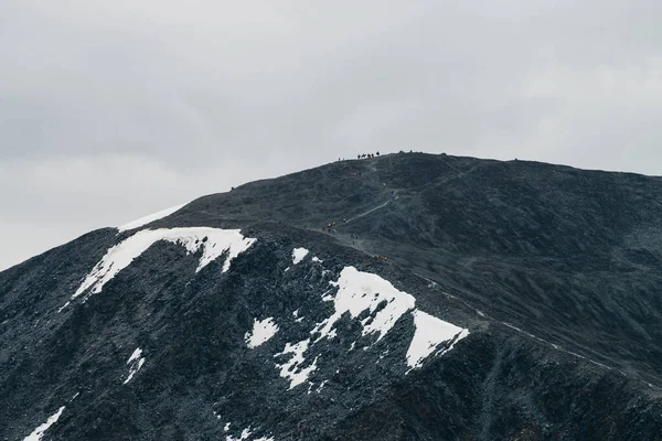 大きな灰色の山の斜面に色のバックパックを持つ多くの人々 灰色の曇りの空の下で巨大な山の中腹に多色の小さな点 観光客との巨大な岩の尾根 山岳観光 高山旅行 — ストック写真