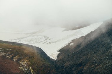 Atmosferik minimalist alp manzarası dağ yamacında, bulutların içinde büyük bir buzulla dağlık vadide. Kayalık vadide alçak bulutlar. Huzurlu dağ manzarası. Karlı dağ yamacının harika manzarası.