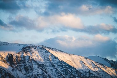 Atmosferik dağlık araziden, gün batımında karlı dağ sırtına. Dağ zirvesinde kar altın ışıkta parlar. Güzel, parlak, karlı bluz. Akşam bulutlu gökyüzü. Altın gün batımı gölgeleriyle harika bir manzara.