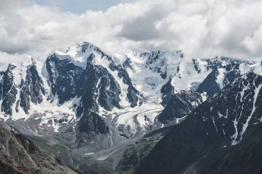 Atmosferik alp manzarası ve dev dağlarda asılı devasa buzullar. Dağ yamacında büyük bir buzulun dili. Karlı dağların arasında alçak bulutlar. Buzdaki çatlaklar. Yüksek irtifada görkemli manzara.