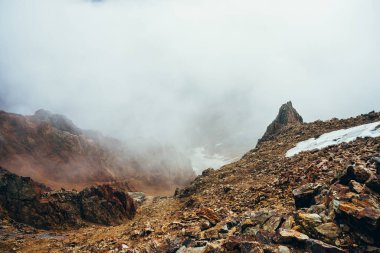 Yüksek kayalık dağ yamacındaki güzel, sivri uçurum, kalın alçak bulutların arasında kar var. Atmosferik minimalist alp manzarası. Uçsuz bucaksız bir uçurumun kenarında büyük bir bulut. Muhteşem dağlık manzara.