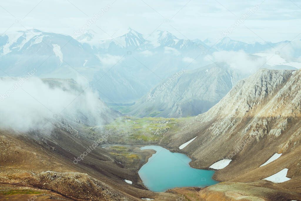 Atmospheric alpine landscape to beautiful glacial lake in highland valley. Big snowy mountains with glacier above mountain lake. Thick low clouds among rocks. Wonderful scenery. Flying over mountains.
