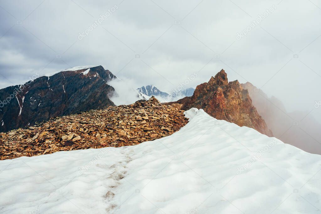 Pointy rocky pinnacle on mountain with snow among thick low clouds. Atmospheric alpine landscape. Footprints in snow on mountain top above thick clouds in abyss. Wonderful highland scenery. Sharp rock