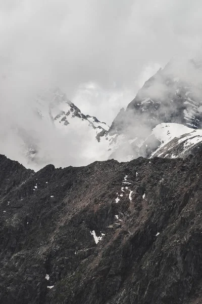 大きな岩の山の壁と低雲の中に雪に覆われたピークと大気中のミニマリストの高山風景 巨大な氷河を持つ巨大な山 雪とクスクス笑う壁 標高の高い雄大な景色 — ストック写真