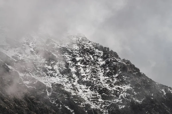 Atmosferik minimalist dağ manzarası ve karlı dağ zirvesi. Kar sınırına yakın alçak bulutlar. Karlı ve sisli kayalar. Sisli Craggy Dağları. Yüksek irtifada görkemli sisli manzara