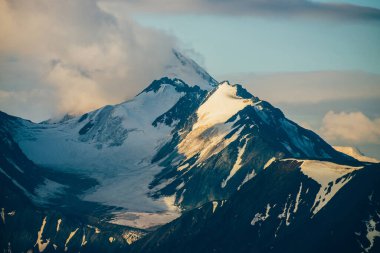 Atmosferik alp manzarası. Altından saatte alçak bulutlar arasında büyük, karlı dağlar. Güneşin doğuşunda dev bir dağ sırasının üzerinde muazzam bir buzulla harika bir dağ manzarası. Büyük kayalıklarda parlak kar.