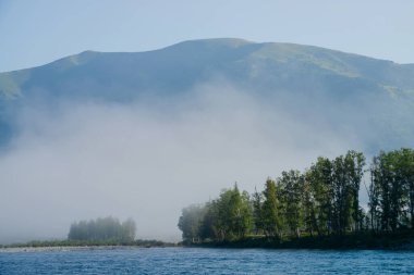 Sis içinde nehrin uzak kıyısındaki büyük dağa harika bir sabah manzarası. Nehrin karşı tarafındaki ağaçların üzerinde güzel bir sis var. Dağlarda hoş bir sabah tazeliğinin huzurlu manzarası. İlham verici manzara.