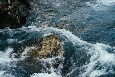Dağ nehrinin masmavi sularında büyük taşlar. Dağ deresinin turkuaz suyunda büyük kaya kütleleri olan mavi bir arka plan. Tam kapasite deniz sörfü. Taşların arasında okyanus sörfü. Gelgitin arkaplanı.