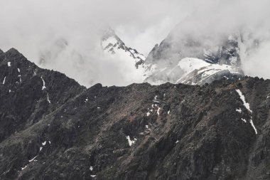 Atmosferik minimalist dağ manzarası. Büyük kayalık bir dağ duvarı ve alçak bulutlarda karla kaplı bir tepe. Devasa buzullu büyük dağlar. Karlı, kayalık bir duvar. Yüksek irtifada görkemli manzara.