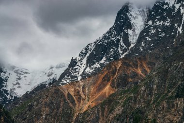 Büyük kayalıkları ve kötü bulutlu havada buzulları olan kasvetli, muhteşem bir manzara. Hafif fırtınalı bulutlar karlı dağın tepesine dokunur. Dağlar yüzünden tehlike fırtınası geliyor. Kasvetli bulutlu atmosferik manzara