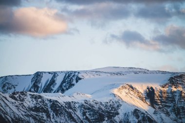 Atmosferik dağlık araziden, gün batımında karlı dağ sırtına. Dağ zirvesinde kar altın ışıkta parlar. Güzel, parlak, karlı bluz. Akşam bulutlu gökyüzü. Altın gün batımı gölgeleriyle harika bir manzara.