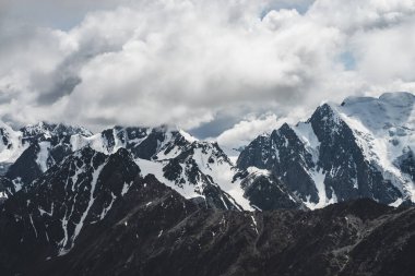 Atmosferik minimalist alp manzarası. Bulutlu gökyüzünün altındaki büyük dağda asılı büyük bir buzulla. Buzdaki çatlaklar. Karla kaplı alçak bulutlar büyük dağ sıraları. Yüksek irtifada görkemli manzara.
