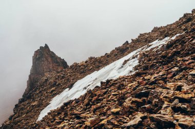 Yüksek kayalık dağ yamacındaki güzel, sivri uçurum, kalın alçak bulutların arasında kar var. Atmosferik minimalist alp manzarası. Uçsuz bucaksız bir uçurumun kenarında büyük bir bulut. Muhteşem dağlık manzara.