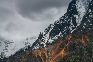Büyük kayalıkları ve kötü bulutlu havada buzulları olan kasvetli, muhteşem bir manzara. Hafif fırtınalı bulutlar karlı dağın tepesine dokunur. Dağlar yüzünden tehlike fırtınası geliyor. Kasvetli bulutlu atmosferik manzara