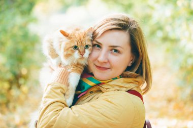 Büyük kırmızı kedi dışarıdaki kızın elinde. Güz renklerinde kızıl kedili güzel bir kadın portresi. İki tembel surat. Yeşil turuncu bokeh 'in arka planında tüylü bir hayvan var. Büyük kedi gözleri yakın çekim..