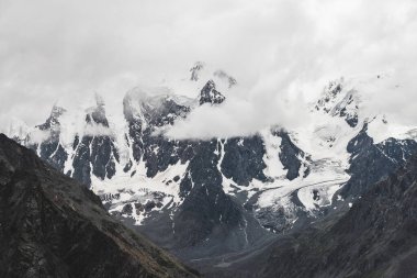 Atmosferik alp manzarası ve dev dağlarda asılı devasa buzullar. Dağ yamacında büyük bir buzulun dili. Karlı dağların arasında alçak bulutlar. Buzdaki çatlaklar. Yüksek irtifada görkemli manzara.