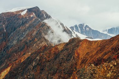 Atmosferik alp manzarası. Kırmızı kayalıklarda alçak bulut var. Büyük turuncu kayalara ve gün doğumunda buzullu dev karlı dağlara güzel bir manzara. Harika bir dağ manzarası. Kayalık duvar.