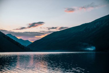 Canlı şafak vakti gökyüzü, gün doğumunda dağ siluetlerinin yakınındaki saf alp gölüne yansıyor. Dağ gölünün sakin suyuyla renkli bir manzara. Güneşli bir sabahta su dalgalanması ile güzel, sakin bir manzara.