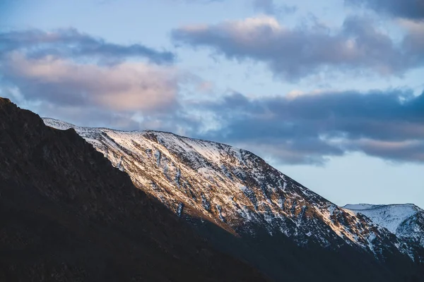 Atmospheric alpine landscape to snowy mountain ridge in sunset. Snow shines in golden light on mountain peak. Beautiful shiny snowy top. Evening cloudy sky. Wonderful scenery in gold sunset shades.