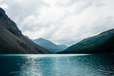 Yağmurlu havada dev dağların arasındaki engin dağ gölüne dramatik bir manzara. Çam ve karaçam yamaçta, gök mavisi suyun yanında. Turkuaz alp gölüyle kaplı bulutlu bir yer. Atmosferik dağ manzarası.