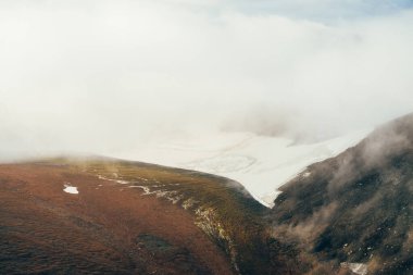 Atmosferik minimalist dağ manzarasından dağ yamacındaki geniş buzullu dağ vadisine kadar. Kayalık vadide alçak bulutlar. Huzurlu dağ manzarası. Karlı dağ yamacının harika manzarası