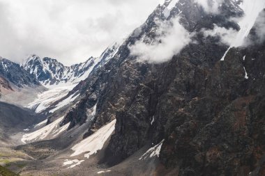 Atmosferik minimalist alp manzarası dev dağlarda asılı devasa bir buzulla. Dağ yamacında büyük bir buzulun dili. Karlı dağların üzerinde alçak bulutlar. Yüksek irtifada görkemli manzara.