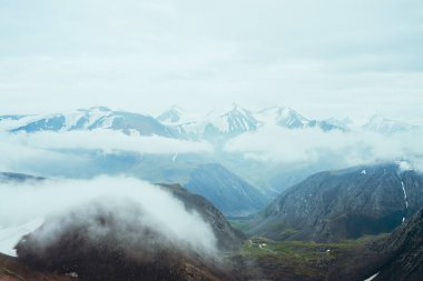 Güzel atmosferik dağlık arazi buzullu karlı dağlara. Küçük nehri olan yeşil vadideki kayaların arasında alçak bulutlar. Harika bir dağ manzarası. Bulutların üzerinde uçarak.