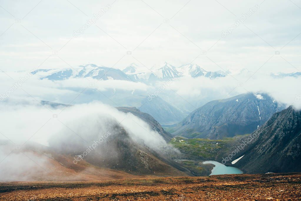 Atmospheric alpine landscape to glacial lake in highland valley. View from stony slope to big snowy mountains with glacier above mountain lake. Thick low clouds among rockies. Wonderful scenery.
