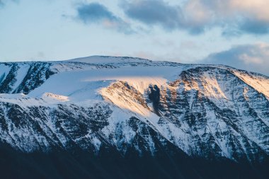 Atmosferik dağlık araziden, gün batımında karlı dağ sırtına. Dağ zirvesinde kar altın ışıkta parlar. Güzel, parlak, karlı bluz. Akşam bulutlu gökyüzü. Altın gün batımı gölgeleriyle harika bir manzara.