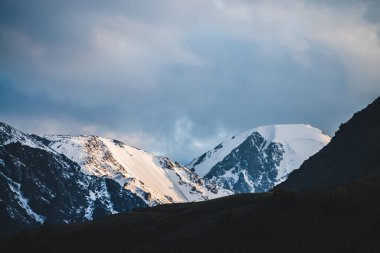 Atmosferik dağ manzarası, gün batımında karlı dağ sırtı ve orman tepeleri. Dağ zirvesinde kar altın ışıkta parlar. Güzel, parlak, karlı bluzu olan harika bir manzara. Akşam bulutlu gökyüzü.