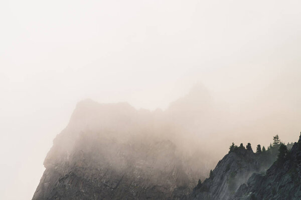 Dramatic bleak fog among giant rocky mountains. Ghostly atmospheric view to big cliff in cloudy sky. Low clouds and beautiful rockies. Minimalist scenery in mysterious place at early foggy morning.