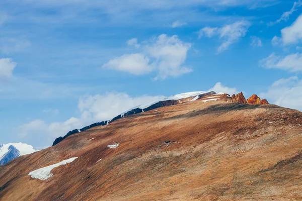 Awesome vivid stony hill with snow in sunlight. Colorful sunny highland landscape with mountain with red crags in top under blue sky. Wonderful alpine scenery with rocks in red orange brown colors.