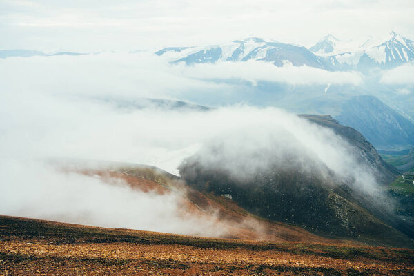 Атмосферный альпийский пейзаж гигантскому низкому облаку над скалистыми горами. Большие густые облака над высокогорной долиной. Прекрасный пейзаж на большой высоте. Вид со склона на заснеженный горный хребет с вершины.