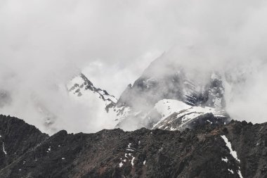 Atmosferik minimalist dağ manzarası. Büyük kayalık bir dağ duvarı ve alçak bulutlarda karla kaplı bir tepe. Devasa buzullu büyük dağlar. Karlı, kayalık bir duvar. Yüksek irtifada görkemli manzara.