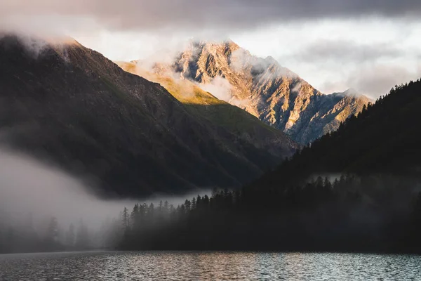 High gold mountain peak and many low clouds above mountain lake at sunrise. Dense fog above water and forest in golden hour. Atmospheric highland landscape at early morning. Alpine relaxing scenery.