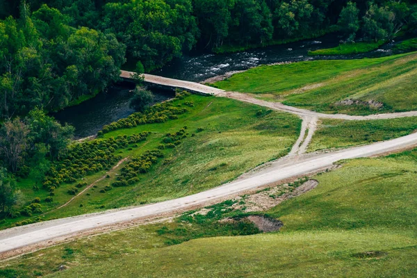 Scenic aerial view to dirt road and wood bridge through mountain river in grove. Atmospheric vivid landscape of countryside with river among trees. Beautiful colorful scenery with road along creek.