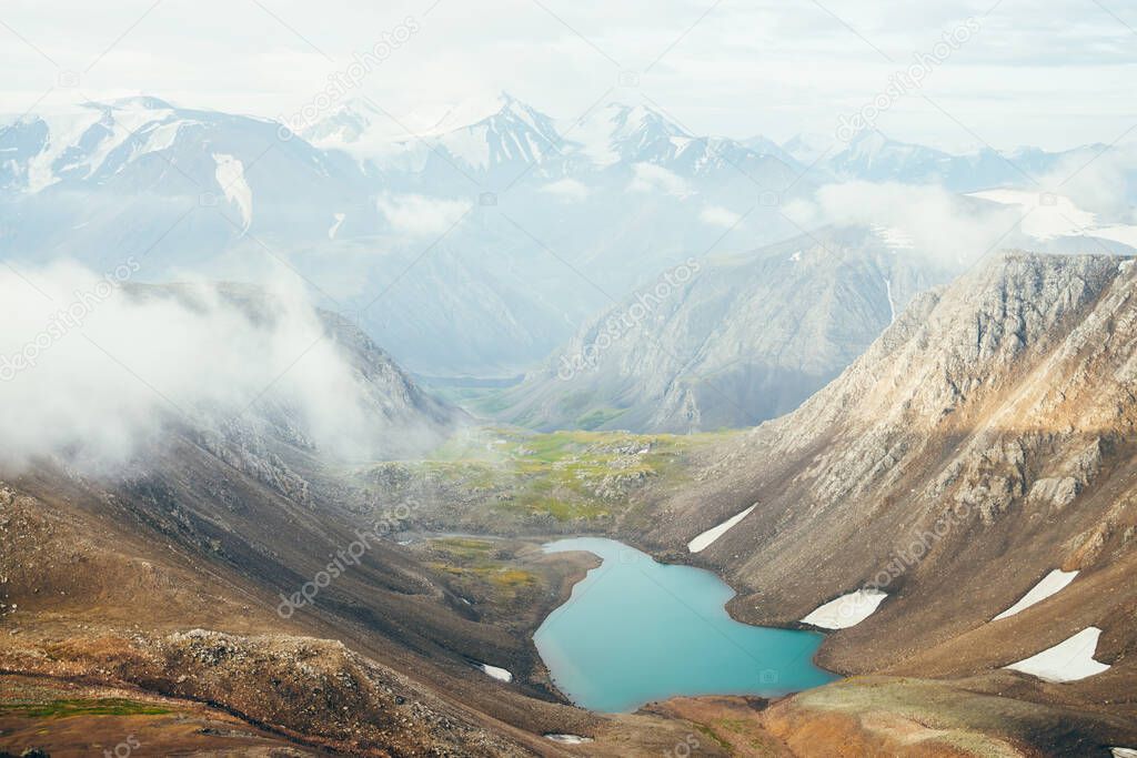 Atmospheric alpine landscape to beautiful glacial lake in highland valley. Big snowy mountains with glacier above mountain lake. Thick low clouds among rocks. Wonderful scenery. Flying over mountains.