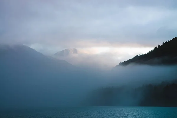Beautiful mountain lake among huge mountains and forest in mist in golden hour. Sun shines through dense low clouds. Big rock glitters at sunrise. Alpine relaxing scenery with fog in pastel tones.