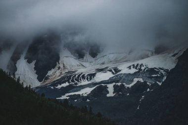 Karanlık bir akşamda alçak bulutların arasındaki dev karlı dağ duvarına kasvetli dramatik bir manzara. Yoğun siste büyük karlı kayalıkları olan kasvetli bir dağ manzarası. Atmosferik sisli hava ve ormanın arkasında büyük bir buzul var..