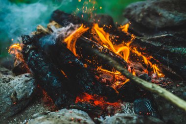 Yanan odun odunları yakın plan ateşte yanmış. Atmosferik sıcak arka plan, turuncu kamp ateşi ve mavi duman. Şenlik ateşinin tam kare görüntüsü. Közlerin güzel kasırgası ve küller havada