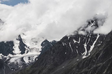 Atmosferik alp manzarası ve dev dağlarda asılı devasa buzullar. Dağ yamacında büyük bir buzulun dili. Karlı dağların arasında alçak bulutlar. Buzdaki çatlaklar. Yüksek irtifada görkemli manzara.