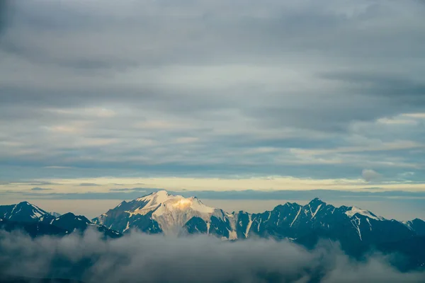 黄金の時間で低雲の間に大きな雪の山と大気高山の風景 日の出の巨大な山脈の巨大な氷河と素晴らしい高原の風景 大きな岩の上に輝く雪 — ストック写真