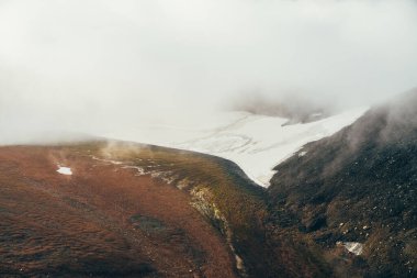 Atmosferik minimalist dağ manzarasından dağ yamacındaki geniş buzullu dağ vadisine kadar. Kayalık vadide alçak bulutlar. Huzurlu dağ manzarası. Karlı dağ yamacının harika manzarası