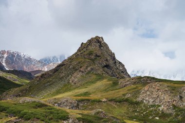 Atmosferik dağ manzarası. Güzel bir tepe kayası ve karlı dağların arasındaki yeşil vadide alçak bulutlar. Yeşil tepenin tepesine ve bulutlu gökyüzünün altında karla kaplı sıradağlara müthiş bir manzara..